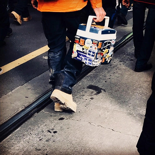 worker walking down a street carrying a lunchbox covered in stickers